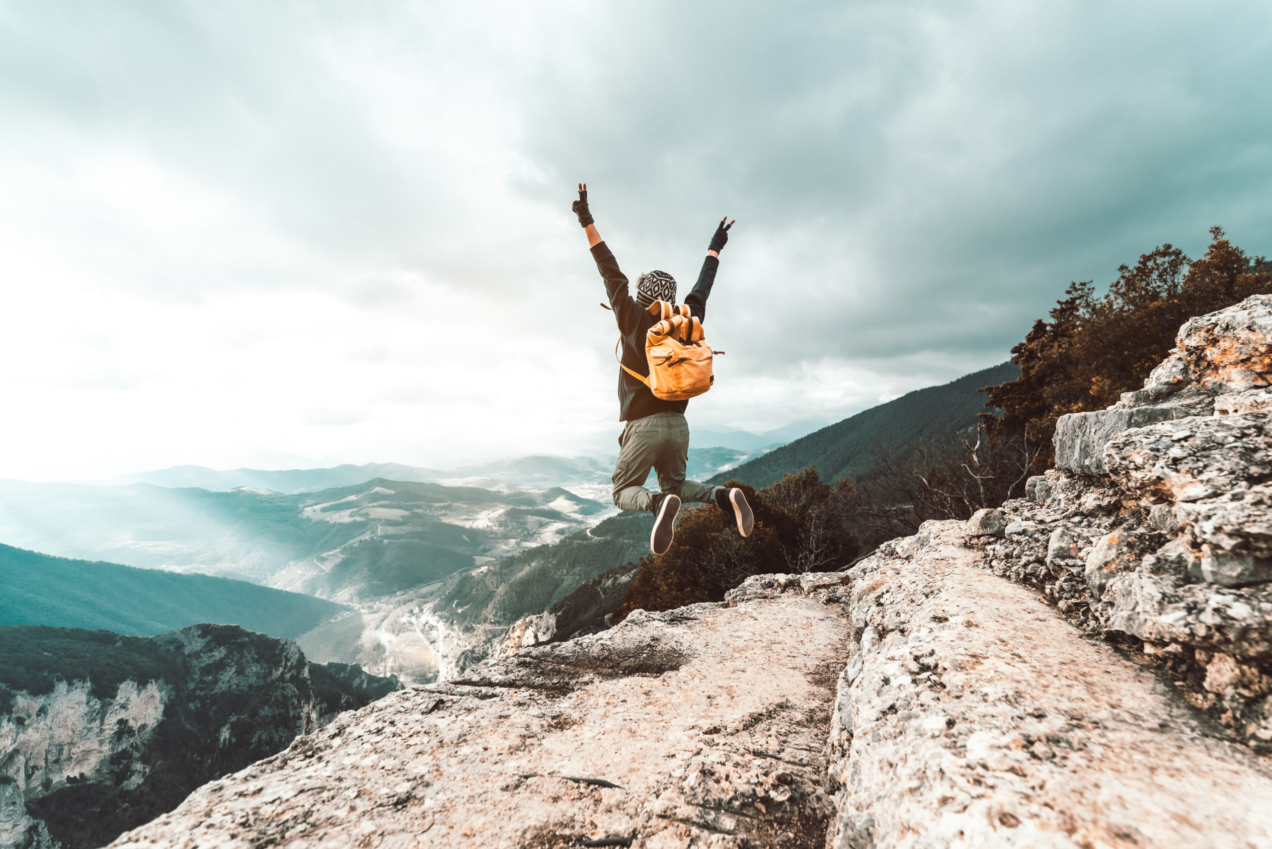 Young man traveler jumps on a background of mountain raising arms to the sky